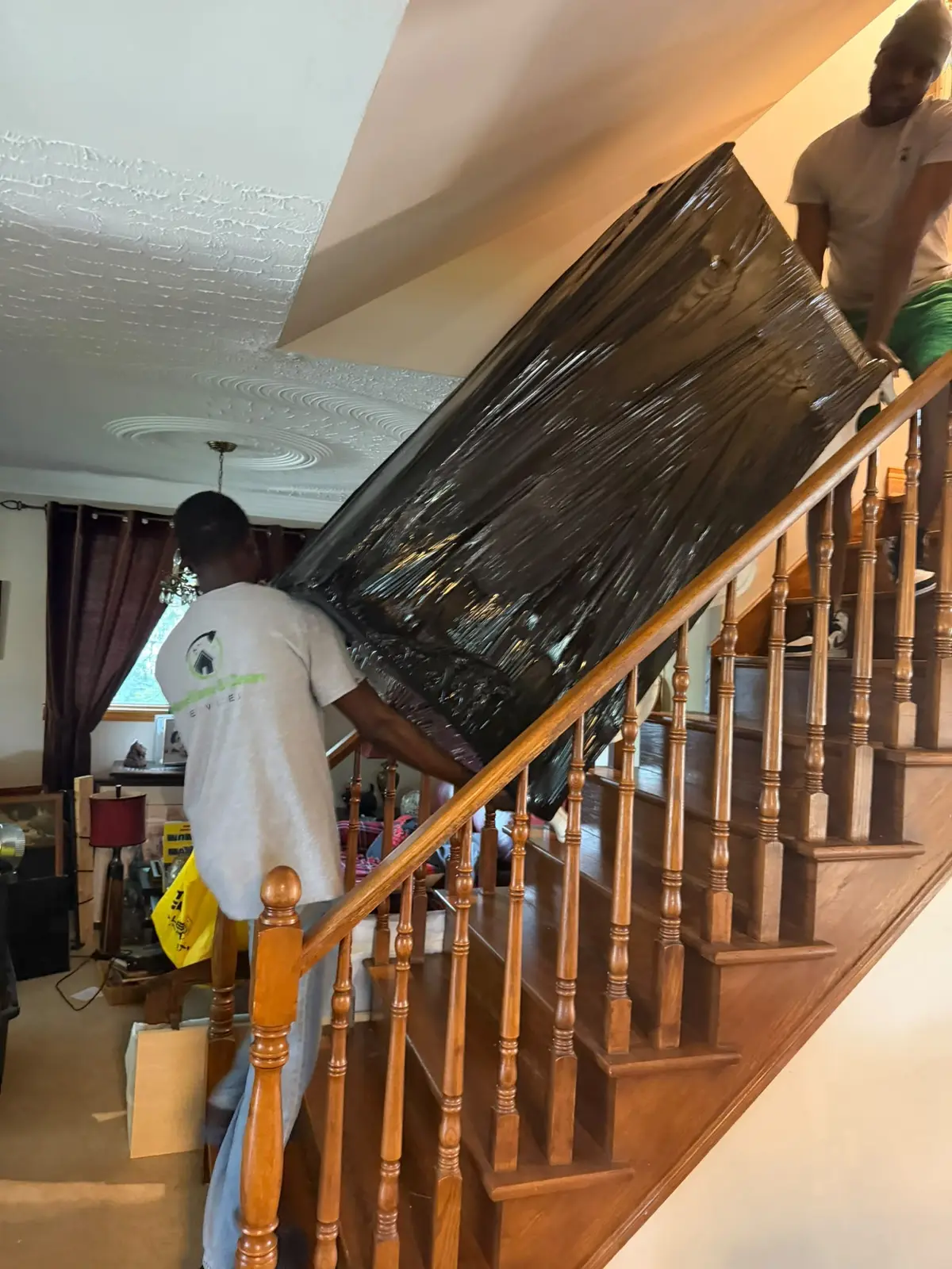 Two movers carrying plastic-wrapped furniture down a staircase